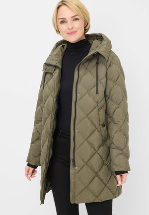 Woman wearing a long olive green quilted jacket with a hood and black zipper, paired with a black turtleneck and black pants.