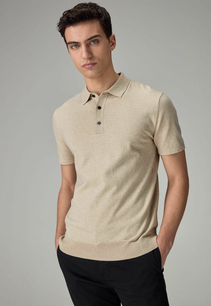 Beige-paitainen polo-paita, jossa on teksturoitu pinta, lyhyet hihat, huomaamaton kaulus ja kolme mustaa napin yksityiskohtaa, yhdistettynä tummiin housuihin.