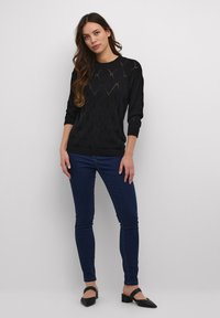 Maglione nero lavorato a maglia con motivi a diamante, scollatura rotonda e maniche a 3/4, abbinato a jeans skinny blu scuro e mule neri.