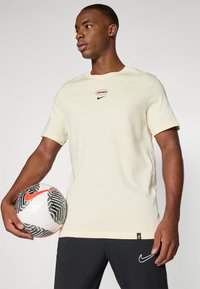 Krémové tričko s krátkým rukávem má kulatý výstřih a červené logo "allmax" nad černým Nike swoosh. Černé sportovní šortky dokončují celý outfit.