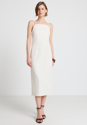 Donna in piedi che indossa un vestito midi bianco senza maniche con bretelle sottili, tacchi scuri con cinturino alla caviglia e un braccialetto marrone al polso destro.