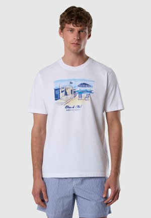 Jeune homme portant un t-shirt blanc avec une scène de plage bleue et le texte « Beach Club North Sails », associé à un short rayé bleu et blanc.