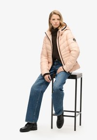 Giacca puff rosa chiaro con cappuccio, dolcevita marrone a maglia, jeans blu e stivali neri con lacci, seduto su uno sgabello di metallo nero.