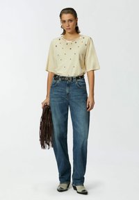 Beživa košulja kratkih rukava s srebrnim ukrasima, uparena s plavim trapericama, crnim remenom sa srebrnim detaljima i sivom obućom.