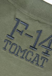 Tkanina w kolorze oliwkowym z wyszytym tekstem "F-14 TOMCAT" w granatowym kolorze. Tekst jest pogrubiony i teksturowany na tle o gładkiej fakturze.