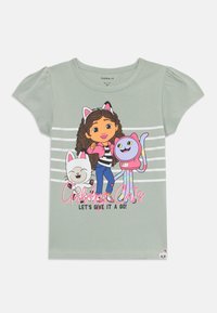 T-shirt de algodão verde claro com um personagem de cabelo castanho encaracolado, orelhas de gato e dois gatos de desenho animado. O texto diz "Gatos Murmuradores! Vamos experimentar!"