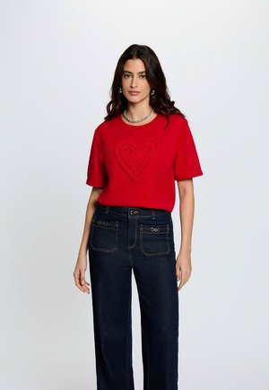 Mujer con una camiseta roja de manga corta con un diseño de corazón texturizado y vaqueros azules oscuros de talle alto, de pie contra un fondo liso.