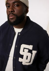 Dunkelblaue Fleece-Varisty-Jacke mit einem weißen "SU"-Logo auf der Brust, ausgestattet mit silbernen Druckknöpfen und einem klassischen Kragen-Design.