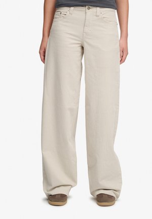 Personne portant un pantalon beige ample, un haut gris foncé et des chaussures marron, debout devant un fond blanc uni.