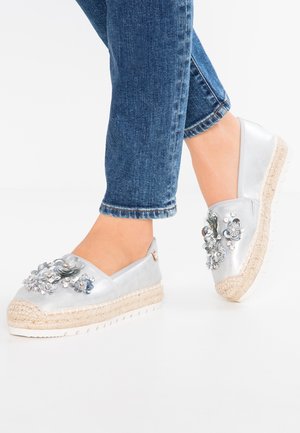 Feet wearing silver slip-on shoes with floral and rhinestone embellishments, paired with blue denim jeans against a white background.