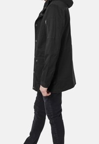 Giacca nera con cappuccio, realizzata in tessuto resistente; presenta bottoni a pressione, tasche laterali e una tasca con zip sul braccio. Design moderno e dalla vestibilità rilassata.