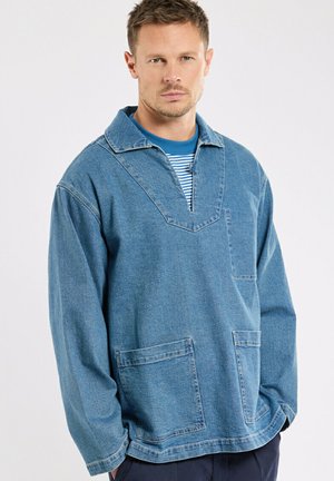 Homme portant un pull en denim ample avec de grandes poches avant par-dessus une chemise à rayures bleues, posant devant un fond uni.