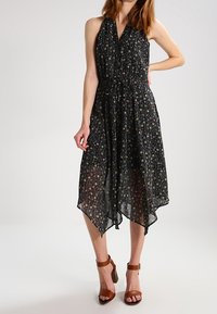 Czarna sukienka w kwiaty z dekoltem na halter, z asymetrycznym dołem i elastyczną talią, z delikatnym wzorem kwiatowym w różnych kolorach. Brązowe sandały na obcasie.