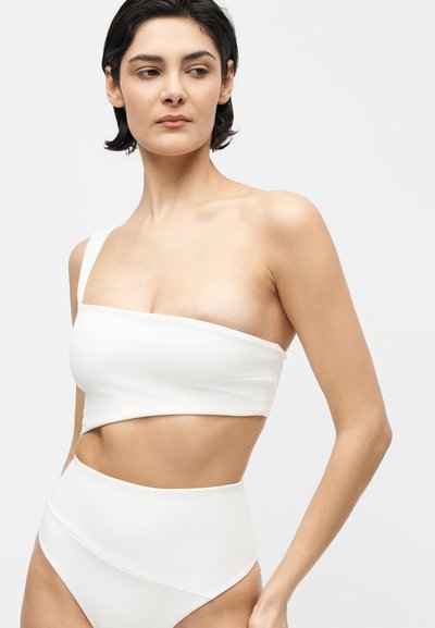 Γυναίκα που φοράει λευκό ασύμμετρο μπικίνι με έναν ώμο και μαγιό με ψηλή μέση, στέκεται μπροστά σε απλό ανοιχτό φόντο.