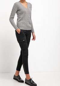 Harmaa kaapelikudottu villapusero V-kauluksella, yhdistettynä mustiin cargo-tyylisiin housuihin, joissa on sivutaskut ja vetoketjut, käytetty kiiltävien mustien kenkien kanssa.