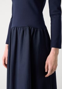 Laivastonsininen mekko, jossa on pitkät hihat; vartalonmyötäinen yläosa siirtyy laskostettuun hameeseen, jossa on sileä rakenne, korostaen virtaavaa muotoilua.