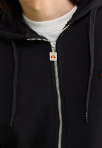 Czarna bluza z kapturem na zamek wykonana z miękkiego materiału. Posiada srebrny zamek błyskawiczny oraz małą, kolorową naszywkę z logo przy szyi. Model z kapturem.