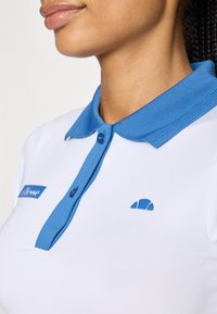 Biela polo košeľa s modrým golierom, so modrým logom a gombíkmi. Hladká, priliehavá tkanina s klasickým dizajnom.