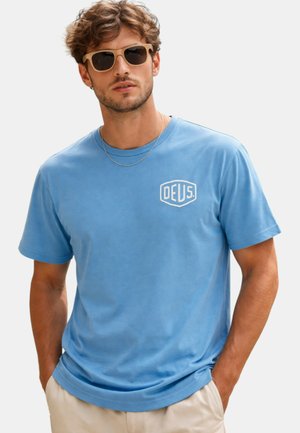 Uomo con capelli ricci che indossa occhiali da sole marroni, una sottile catena d'argento, una maglietta blu Deus e pantaloni beige con le mani in tasca.