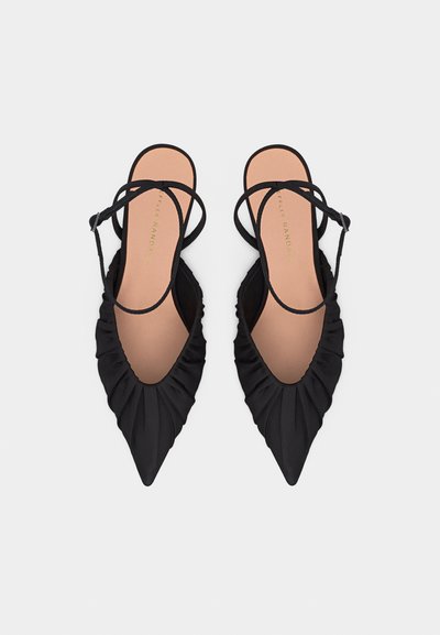 Μαύρα φλατ παπούτσια με μυτερή μύτη, ρίγες λεπτομέρειες και λεπτά λουράκια στον αστράγαλο σε ανοιχτό φόντο.