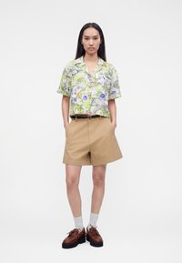 Femme portant une chemise à manches courtes à motif floral, un short beige taille haute avec ceinture, des chaussettes blanches et des chaussures à plateforme marron, debout les mains dans les poches.