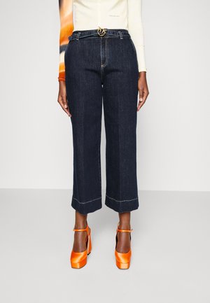 Person wearing dark wide-leg cropped jeans with a black belt, multicolored long-sleeve top, and bright orange platform heels.