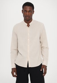 Camicia beige chiaro a maniche lunghe con sottili righe verticali, colletto alla cinese e chiusura frontale con bottoni. Il tessuto sembra tessuto e morbido.