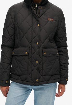 Veste matelassée noire avec boutons-pression à l'avant, deux poches à rabat et un col zippé portée sur un jean bleu clair.