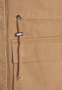 Khaki stof med en tekstureret overflade, prydet med en metaløje og snøre med detaljer på en lomme. Enkel design og neutral farve.