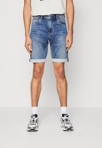 Vaalean siniset farkkushortsit, joissa on kulunut design, käänteet, viisi taskua ja vetoketju, yhdistettynä valkoisiin sukkiin ja tennareihin.