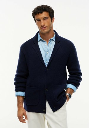 Hombre que lleva un cárdigan azul marino abotonado sobre una camisa azul claro con mangas remangadas y pantalones blancos, posando con una mano en el bolsillo.