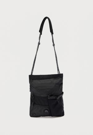 Sac bandoulière noir avec sangle en corde réglable, poche zippée à l'avant, pochette en filet et boucle en tissu sur un fond uni.