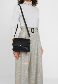 Žena v světle šedém dlouhém rukávu a béžových kostkovaných kalhotách s vysokým páskem a černým opaskem, nese černou koženou kabelku přes rameno.