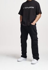 T-shirt noir surdimensionné avec l'inscription "2Y STUDIOS" en blanc ; associé à un pantalon cargo noir avec plusieurs poches ; baskets blanches.