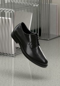 Chaussure habillée en cuir noir avec un bout rond, un design à lacets, une texture lisse et des accents cousus sur une surface réfléchissante.