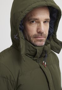 Olivgrön puffjacka med huva, med en marinblå foder, synlig dragkedja och dragsnoddar med röda accenter. Strukturerad tyg.