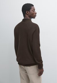 Donkerbruin gebreide trui met een geribbelde structuur, voorzien van een hoge kraag en aansluitende manchetten, gecombineerd met lichtgekleurde corduroy broek.