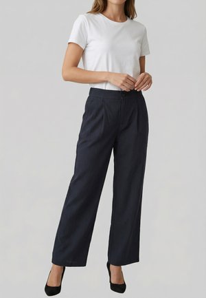 Femme portant un t-shirt blanc à manches courtes, un pantalon noir taille haute à jambes larges, et des talons noirs, debout devant un fond clair uni.