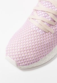 Světle růžový teniskový model s béžovými tkaničkami a fialovým síťovinovým překryvem na svršku, prezentován v detailu na bílém pozadí.