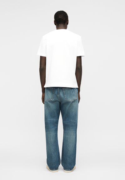 Άτομο με κοντά κατσαρά μαλλιά, που φοράει ένα απλό λευκό μπλουζάκι, μπλε τζιν και ανοιχτόχρωμα παπούτσια, στέκεται με την πλάτη σε ένα απλό φόντο.