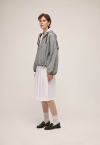 Femme debout portant une veste grise à capuche, une jupe plissée blanche, des chaussettes blanches et des mocassins noirs, sur un fond uni.