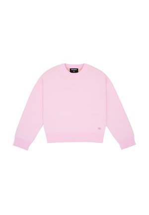 Femme debout, portant un pull rose ample, un short bleu à fines rayures et des chaussures blanches à lacets bleus sur un fond uni clair.