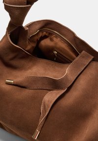 Brun suede håndtaske med en blød tekstur, der har en indvendig lynlåslomme og et enkelt design. Hardwaredetaljer findes på stroppen.