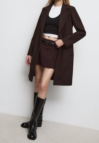 Cappotto lungo marrone sopra un top nero a maniche corte e una gonna plissettata bordeaux, abbinati a stivali alti in pelle nera e una cintura nera.