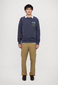 Marineblå sweatshirt med blomsterbroderi, ribbede mansjetter, og en lyseblå skjorte med krage under, sammen med khaki bukser og mørke sko.