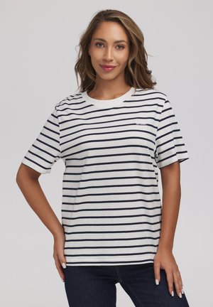 Kobieta z włosami do ramion, ubrana w białą koszulkę z czarnymi poziomymi paskami i ciemne jeansy, uśmiechająca się z jedną ręką na biodrze.