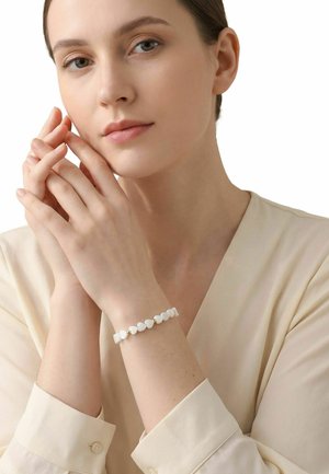 Mujer de piel clara y cabello castaño que lleva una blusa color crema y una pulsera de cuentas blancas en forma de corazón, tocándose suavemente el rostro con ambas manos.