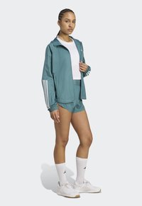 Teal leichter Windbreaker mit Kapuze, weißen Akzenten und elastischen Bündchen; kombiniert mit tealfarbigen Shorts. Weiße Sportschuhe und Socken.