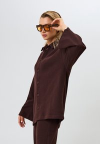 Bruine button-up shirt met een ribbel-textuur, oversized mouwen en een kraagontwerp. Draagt tortoise zonnebril en steil haar.