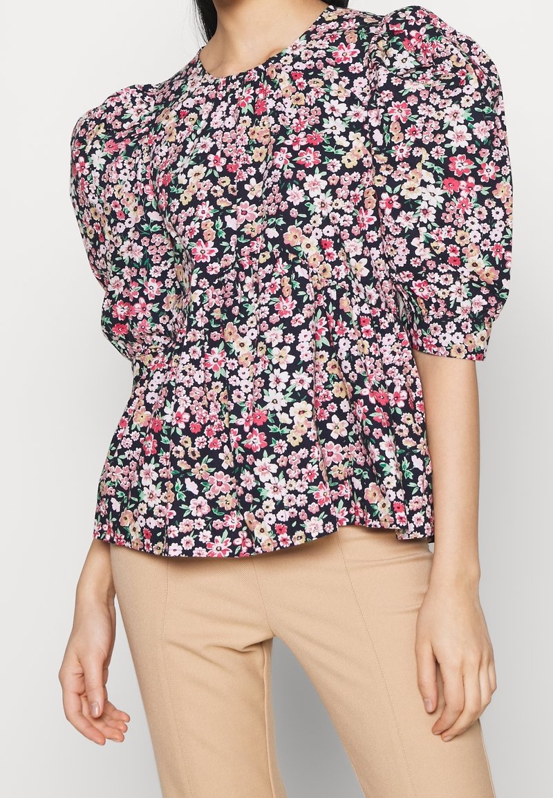 Haut à basque floral en bleu marine foncé, avec des fleurs roses et blanches. Manches bouffantes avec coutures froncées et encolure ronde. Tissu léger.